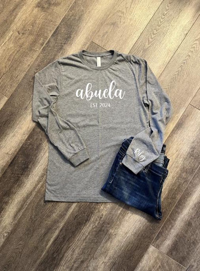 Pu&ograve; includere: Maglietta grigia a maniche lunghe con il testo "abuela est 2024" in bianco. La maglietta &egrave; abbinata a un paio di jeans blu.