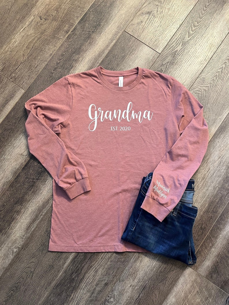 Pu&ograve; includere: Una maglietta a maniche lunghe color rosa polvere con la scritta "Grandma Est. 2020" stampata in bianco sul davanti. La manica sinistra ha la scritta "Maverick Madison" stampata in bianco.