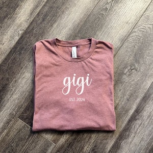 Pu&ograve; includere: Una maglietta color malva con la parola "gigi" scritta in corsivo bianco e il testo "EST. 2024" sotto.