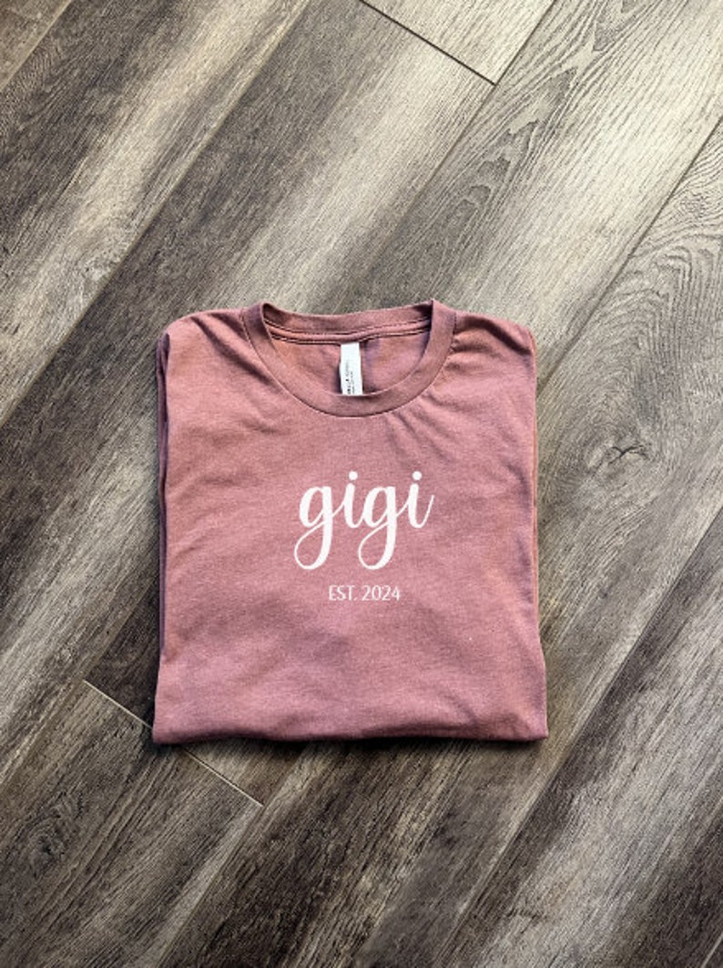 Pu&ograve; includere: Una maglietta color malva con la parola "gigi" scritta in corsivo bianco e il testo "EST. 2024" sotto.