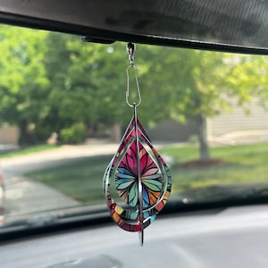 May include: A colorful, teardrop-shaped car charm with a stained-glass design. The charm features a vibrant array of colors including pink, red, blue, green, and yellow. It hangs from a silver-colored clip, adding a decorative touch to the car's interior.
