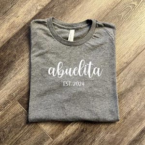 Pu&ograve; includere: Una maglietta grigia con la scritta "abuelita est. 2024" stampata in bianco.