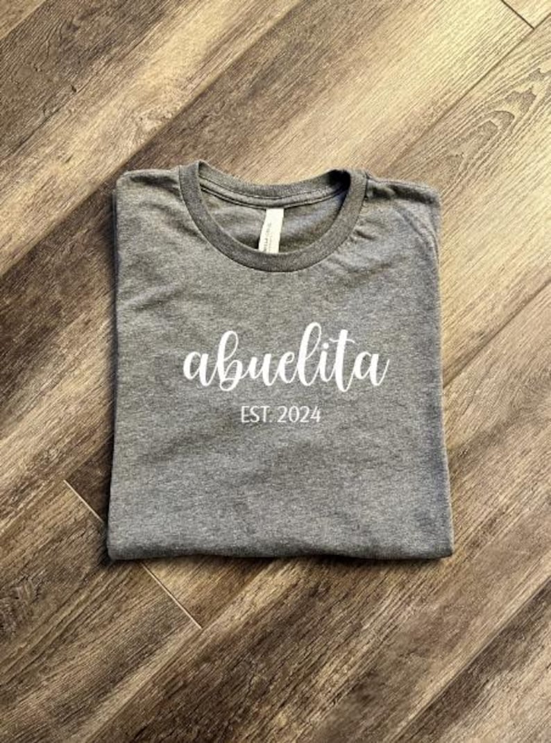 Pu&ograve; includere: Una maglietta grigia con la scritta "abuelita est. 2024" stampata in bianco.