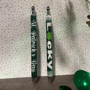 May include: Two green glitter pens with silver accents. One pen says "Lucky" and the other says "St. Patrick's Day".