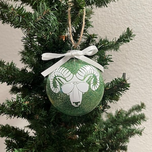 May include: A green glitter Christmas ornament with a white ram head wearing sunglasses. The ornament is hanging from a twine string and a white ribbon bow. The ornament is displayed on a green Christmas tree.