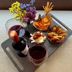 May include: A dark gray wooden serving tray with a bottle of red wine, two wine glasses, and small bowls of snacks. The snacks include chips, cheese, and fried foods. A small vase of colorful flowers sits on the tray.