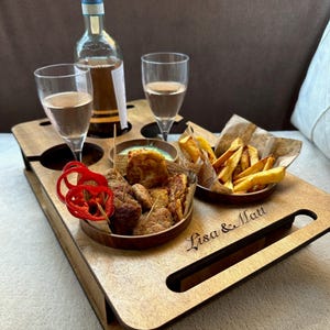 May include: A wooden serving tray with cutouts for two wine glasses and a bottle. The tray holds small bowls of food and a bottle of wine. The tray is engraved with the names "Lisa & Matt".