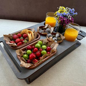 Puede incluir: Una bandeja de madera con varios alimentos. Incluye fresas, gofres, cerezas y pequeñas bolas de chocolate en cuencos de madera. También hay dos vasos de zumo de naranja y un jarrón con flores coloridas.