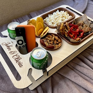 Könnte beinhalten: Ein TV-Tablett aus hellem Holz mit Fächern für Snacks und Getränke. Es hat zwei Getränkehalter, ein Fach für Telefon und Fernbedienung und ist mit Popcorn, Chips, Nüssen und anderen Snacks gefüllt. Die Namen "John & Olivia" sind eingraviert.