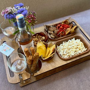 Puede incluir: Una bandeja de madera con una botella de vino, dos copas y pequeños cuencos con aperitivos. Los aperitivos incluyen palomitas de maíz, patatas fritas, nueces y salchichas. También hay un pequeño ramo de flores y una etiqueta de "Gracias".
