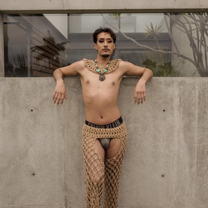 Puede incluir: Un conjunto de crochet beige que consiste en un collar halter con detalles turquesa y pantalones de tejido abierto. El modelo lleva ropa interior Calvin Klein. El conjunto está sobre una pared de hormigón con una ventana.