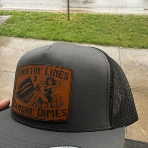 May include: A gray trucker hat with a black mesh back and a brown leather patch. The patch features the text "SHORTIN' LINES & BANGIN' DIMES" with a pin-up girl illustration and dice. The hat has a snapback closure.