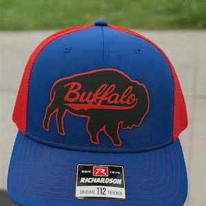 May include: A blue and red trucker cap featuring a black buffalo design and the word "Buffalo" in red script. The cap has a mesh back and a curved peak. The Richardson label is visible.