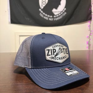 以下が含まれることがあります： ネイビーブルーのトラッカーハット。グレーのメッシュバックとカーブしたつばが特徴です。「CERTIFIED ZIP TIE MECHANIC」と白文字で書かれたパッチが付いています。背景には「POW-MIA YOU ARE NOT FORGOTTEN」と書かれた白黒の旗があります。