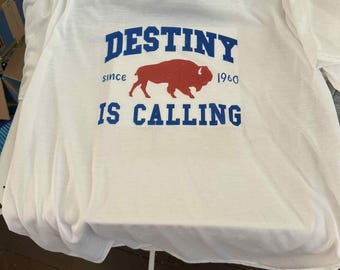 May include: White t-shirt featuring the text "DESTINY IS CALLING" in blue, with a red buffalo graphic. The words "since 1960" are also included. The shirt is made of a soft, comfortable material.