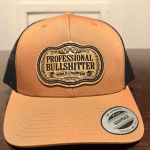 Peut inclure: Casquette de camionneur beige et noire avec un écusson doré portant l'inscription "PROFESSIONAL BULLSHITTER WORLD CHAMPION". La casquette a une visière incurvée et une fermeture à pression. Le texte est en noir.