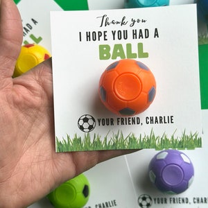 May include: A square white card featuring a small orange soccer ball toy attached to the center. The card displays the text Thank you I HOPE YOU HAD A BALL in black and green, a soccer ball icon, and the text YOUR FRIEND, CHARLIE above green grass.
