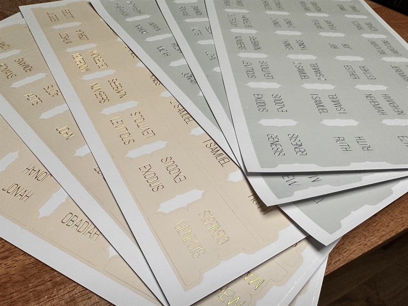 May include: A set of beige and gray book labels with gold lettering. The labels feature the names of books in the Bible, including Genesis, Exodus, Leviticus, Numbers, Deuteronomy, Joshua, Judges, Ruth, 1 Samuel, 2 Samuel, 1 Kings, 2 Kings, 1 Chronicles, 2 Chronicles, Ezra, Nehemiah, Esther, Job, Psalms, Proverbs, Ecclesiastes, Song of Solomon, Isaiah, Jeremiah, Lamentations, Ezekiel, Daniel, Hosea, Joel, Amos, Obadiah, Jonah, Micah, Nahum, Habakkuk, Zephaniah, Haggai, Zechariah, Malachi, Matthew, Mark, Luke, John, Acts, Romans, 1 Corinthians, 2 Corinthians, Galatians, Ephesians, Philippians, Colossians, 1 Thessalonians, 2 Thessalonians, 1 Timothy, 2 Timothy, Titus, Philemon, Hebrews, James, 1 Peter, 2 Peter, 1 John, 2 John, 3 John, Jude, and Revelation.