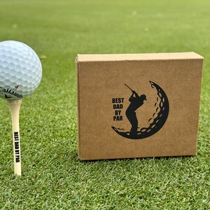 Puede incluir: Una pelota de golf en un tee con "BEST DAD BY PAR" impreso, junto a una caja marr&oacute;n con un dise&ntilde;o tem&aacute;tico de golf y el mismo texto. Los art&iacute;culos est&aacute;n en un campo de golf verde.