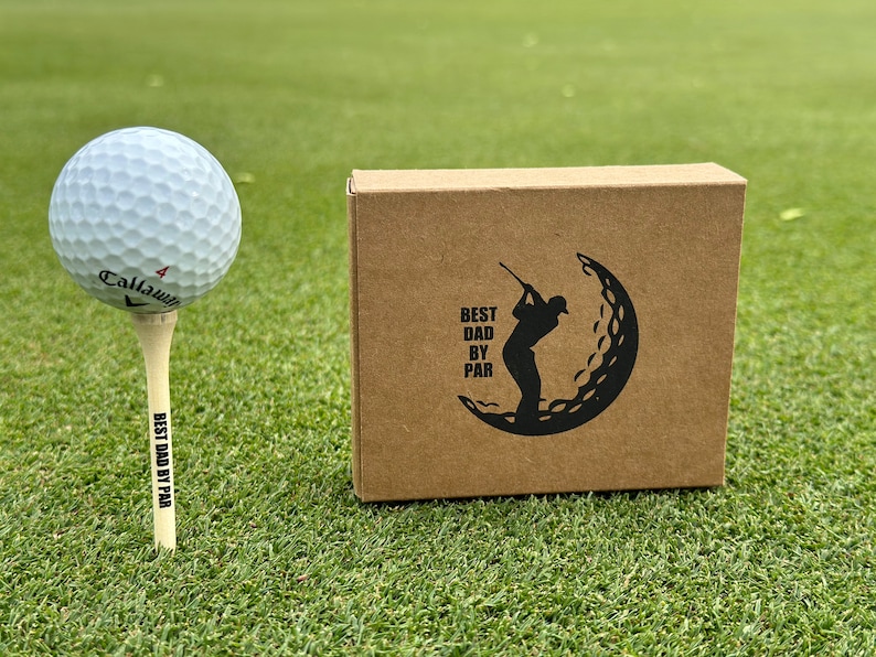 Puede incluir: Una pelota de golf en un tee con "BEST DAD BY PAR" impreso, junto a una caja marr&oacute;n con un dise&ntilde;o tem&aacute;tico de golf y el mismo texto. Los art&iacute;culos est&aacute;n en un campo de golf verde.