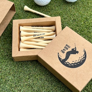 Puede incluir: Una caja de regalo de papel kraft con las palabras "Best Dad by Par" impresas en la tapa y un gr&aacute;fico de pelota de golf. Dentro de la caja hay tees de golf de madera, tambi&eacute;n impresos con "Best Dad by Par". La caja est&aacute; abierta, revelando los tees, y se encuentra sobre una superficie verde.