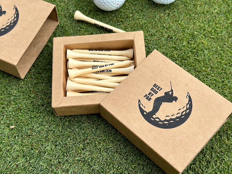 Puede incluir: Una caja de regalo de papel kraft con las palabras "Best Dad by Par" impresas en la tapa y un gr&aacute;fico de pelota de golf. Dentro de la caja hay tees de golf de madera, tambi&eacute;n impresos con "Best Dad by Par". La caja est&aacute; abierta, revelando los tees, y se encuentra sobre una superficie verde.