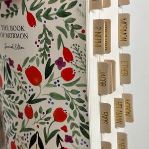 May include: A white journal with a floral pattern and gold tabs labeled with the names of the books of the Book of Mormon. The journal is titled "The Book of Mormon Journal Edition".
