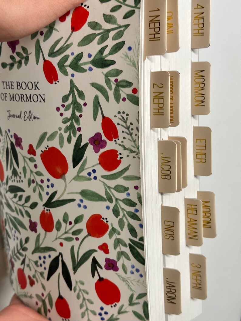 May include: A white journal with a floral pattern and gold tabs labeled with the names of the books of the Book of Mormon. The journal is titled "The Book of Mormon Journal Edition".