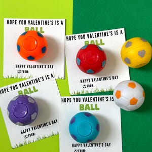 May include: Four Valentine's Day cards with the text "Hope you Valentine's is a ball" and a small soccer ball attached to each card. The soccer balls are orange, red, purple, and blue.