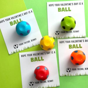 May include: Four Valentine's Day cards with the message "Hope your Valentine's Day is a BALL" and a small soccer ball attached to each card. The soccer balls are blue, green, yellow, and orange.
