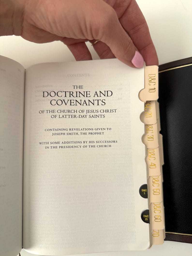 May include: A white book with the title "The Doctrine and Covenants of the Church of Jesus Christ of Latter-day Saints" on the cover. The book has a cream-colored spine with gold lettering and a gold-colored bookmark.