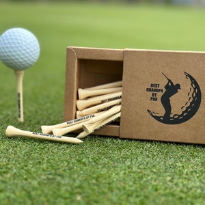 Puede incluir: Una pelota de golf sobre un tee y una caja de tees de golf sobre hierba verde. La caja es marr&oacute;n y tiene un gr&aacute;fico negro de un golfista y el texto "BEST GRANDPA BY PAR". Los tees de golf son de madera y tienen el texto "BEST GRANDPA BY PAR" impreso.