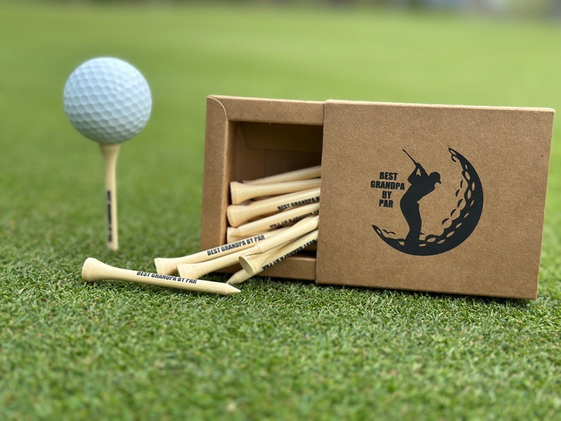 Puede incluir: Una pelota de golf sobre un tee y una caja de tees de golf sobre hierba verde. La caja es marr&oacute;n y tiene un gr&aacute;fico negro de un golfista y el texto "BEST GRANDPA BY PAR". Los tees de golf son de madera y tienen el texto "BEST GRANDPA BY PAR" impreso.