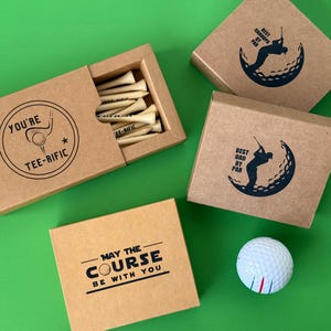 Puede incluir: Un conjunto de regalos con tem&aacute;tica de golf sobre una superficie verde. Incluye una caja de tees de golf, cajas con gr&aacute;ficos y texto relacionados con el golf y una pelota de golf. Las cajas tienen texto que dice "You're Tee-rific", "Best Dad by Par" y "May the Course Be With You".