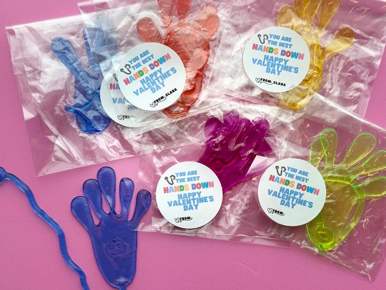 Może przedstawiać: Pięć przezroczystych, plastikowych zabawek antystresowych w kształcie dłoni z kolorowymi naklejkami tęczowymi, na kt&oacute;rych widnieje napis "You are the best hands down Happy Valentine's Day".