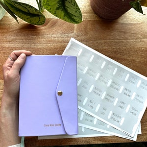 Peut inclure: Un journal en cuir de couleur lavande avec une fermeture à rabat et un bouton doré. Le journal est personnalisé avec le nom "Olivia Mariz Dudley". L'image montre également une feuille d'autocollants avec les noms des livres de la Bible.