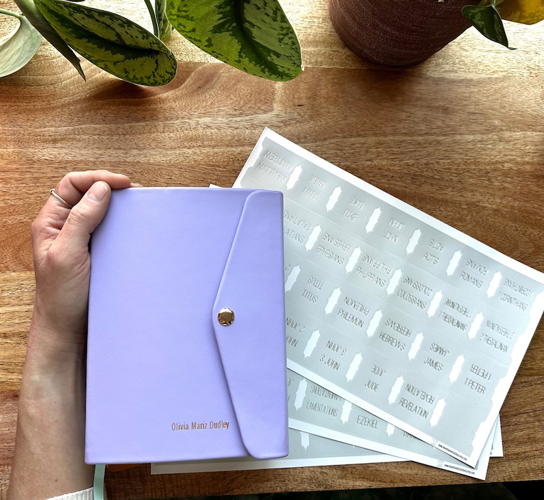 May include: A lavender-colored leather journal with a flap closure and a gold button. The journal is personalized with the name "Olivia Mariz Dudley". The image also shows a set of stickers with the names of books of the Bible.