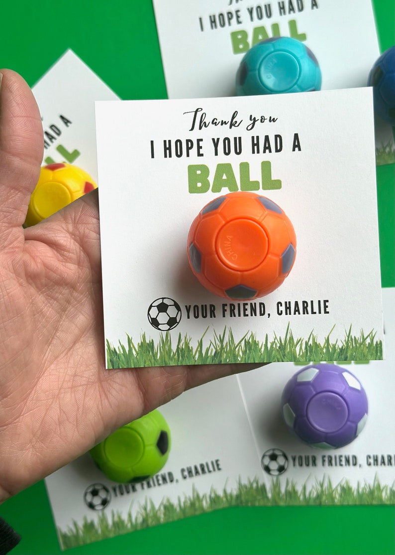 May include: Thank you I hope you had a ball. A small orange soccer ball with black accents is attached to a white card with black text. The card has a small black and white soccer ball graphic at the bottom. The text on the card reads "Thank you I hope you had a ball. Your friend, Charlie."