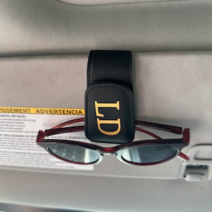 May include: Black leather sunglass holder with gold lettering that says "LD" attached to a car visor. Brown sunglasses are clipped to the holder.