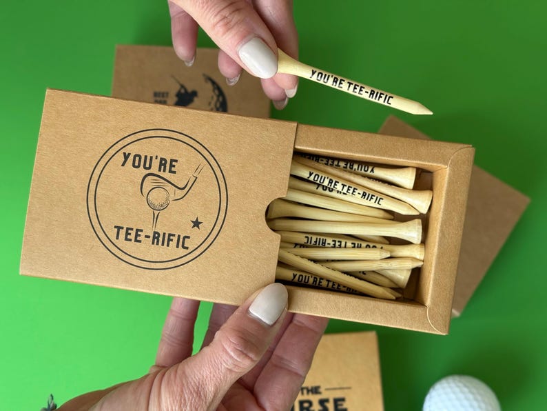 Puede incluir: Una caja de papel kraft con tees de golf de madera, impresos con "YOU'RE TEE-RIFIC". La caja tiene un gr&aacute;fico circular con una pelota y un palo de golf. Se ve una pelota de golf.