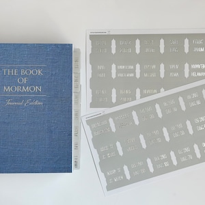 May include: A blue book titled "The Book of Mormon, Journal Edition" with a set of grey and white book dividers labeled with the names of the books in the Book of Mormon and chapter numbers.