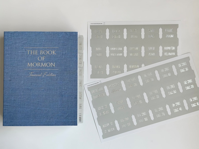 May include: A blue book titled "The Book of Mormon, Journal Edition" with a set of grey and white book dividers labeled with the names of the books in the Book of Mormon and chapter numbers.