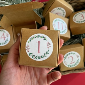 May include: A small, brown cardboard box with a white sticker featuring the number "1" in a red font, surrounded by a green and red wreath. Other boxes with numbers 2, 3, 4, and 11 are visible in the background, suggesting an advent calendar.