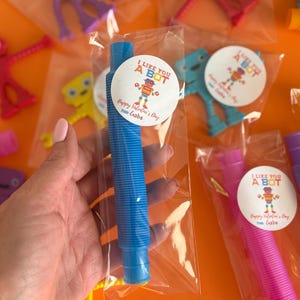 Może przedstawiać: Niebieska plastikowa zabawka ze sprężystym wzorem spiralnym, zapakowana w przezroczystą plastikową torebkę z naklejką walentynkową z napisem "I Like You A Bot. Happy Valentine's Day from Gabe."