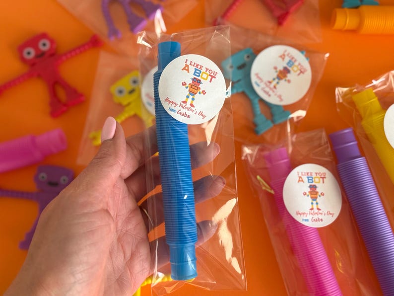 Może przedstawiać: Niebieska plastikowa zabawka ze sprężystym wzorem spiralnym, zapakowana w przezroczystą plastikową torebkę z naklejką walentynkową z napisem "I Like You A Bot. Happy Valentine's Day from Gabe."