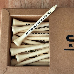 Puede incluir: Una caja de tees de golf de madera natural. Un tee est&aacute; en &aacute;ngulo sobre los dem&aacute;s, mostrando el texto "MAY THE COURSE BE WITH YOU" en negro. La caja es de cart&oacute;n marr&oacute;n.