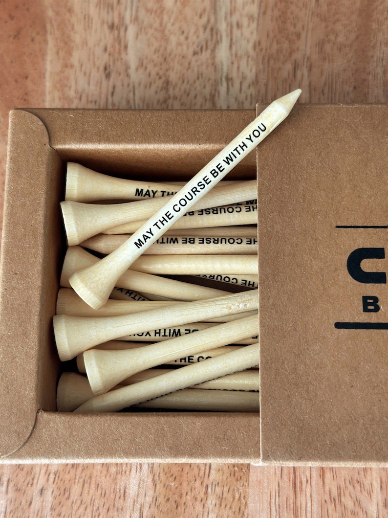 Puede incluir: Una caja de tees de golf de madera natural. Un tee est&aacute; en &aacute;ngulo sobre los dem&aacute;s, mostrando el texto "MAY THE COURSE BE WITH YOU" en negro. La caja es de cart&oacute;n marr&oacute;n.