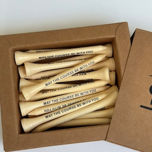 Puede incluir: Una caja de tees de golf de madera clara. Cada tee tiene la frase "MAY THE COURSE BE WITH YOU" impresa en negro. La caja es de cart&oacute;n marr&oacute;n.