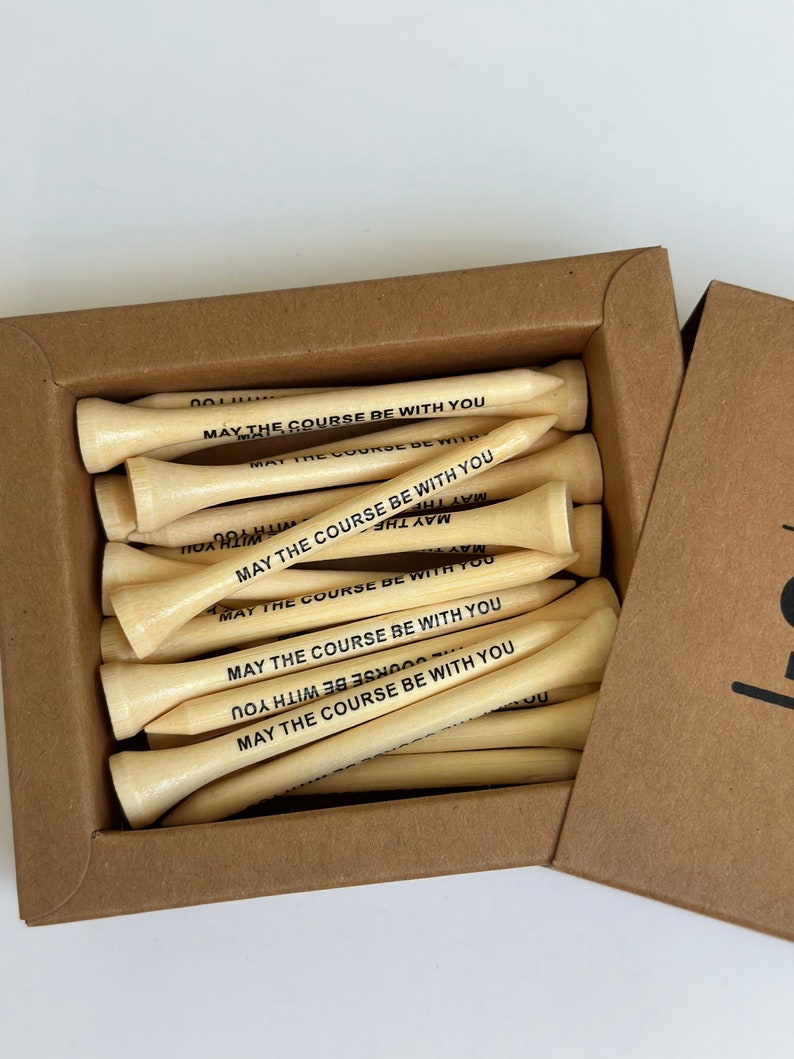 Puede incluir: Una caja de tees de golf de madera clara. Cada tee tiene la frase "MAY THE COURSE BE WITH YOU" impresa en negro. La caja es de cart&oacute;n marr&oacute;n.