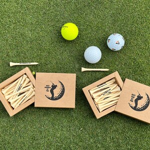 Puede incluir: Juego de regalo con tem&aacute;tica de golf que incluye pelotas de golf, tees de madera y cajas con la silueta de un golfista. Las cajas y los tees tienen el texto "BEST GRANDPA BY PAR". Las pelotas son blancas, amarillas y estampadas, sobre una superficie verde.